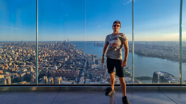 A man in a cap standing at the observatory deck of The Edge with captivating aerial view of New York City skyline over the Hudson River during the dusk. Endless rows of tall buildings. Travelling