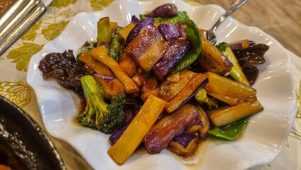 Fried vegetables in Sichuan style, served on a white plate. There are eggplants, broccoli and green...