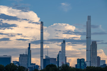 Fototapeta premium Captivating New York urban skyline at dusk with striking and modern skyscrapers reflecting on water seen from Jacqueline Kennedy Oasis Reservoir. The buildings are taller than surrounding clouds.