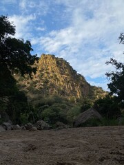 Montaña rocosa de tono amarillo con nubes y arboles, con luz de sol cálida