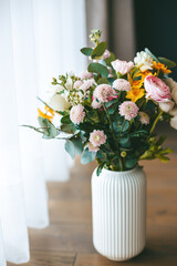 A lively colorful bouquet of various flowers in a white, ribbed vase, set on a wooden floor, with the soft natural light from window
