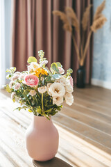 A vibrant bouquet of assorted flowers in a textured pink vase, placed on a wooden floor against the backdrop of elegant pink curtains