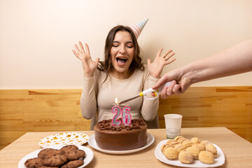 A girl sits in front of a table with a festive cake, on which a candle is lit in the form of the...