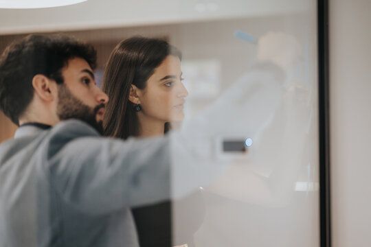 A diverse team of business professionals intensely brainstorming and writing on a glass wall, covered with sticky notes in a bright office setting. - Powered by Adobe