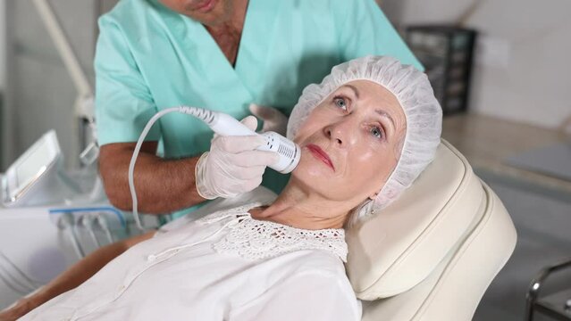 Confident cosmetologist performing anti-aging face hardware therapy procedure for aged female client in clinic of aesthetic medicine