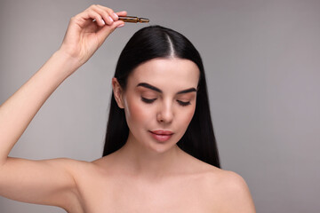 Beautiful young woman using ampoule for hair treatment on grey background