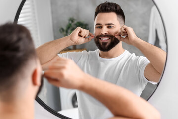 Handsome young man touching mustache near mirror in bathroom