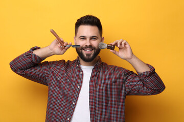 Handsome young man with mustache holding blade and shaving brush on yellow background