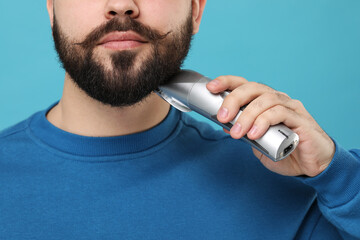 Handsome young man trimming beard on light blue background, closeup