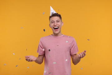 Happy man in party hat throwing confetti on orange background