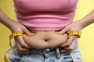 Woman with measuring tape touching belly fat on yellow background, closeup. Overweight problem