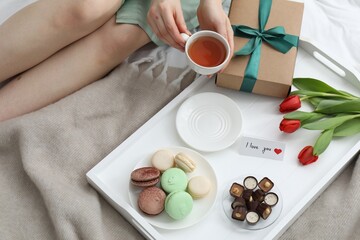Tasty breakfast served in bed. Woman with tea, desserts, gift box, flowers and I Love You card at home, closeup