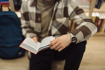 In a warmly lit library, a young man in a stylish sweater is deeply focused on reading an open...