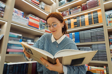 A young woman with glasses reads an interesting classic novel in a well-stocked cozy bookstore,...