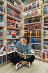 A young woman in glasses is absorbed in reading a large book amidst rows of colorful bookshelves in...