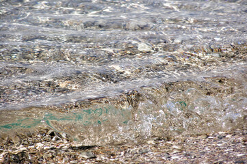 Crystal clear water rippling on rocky shoreline. Transparent water waves gently lapping over a bed...