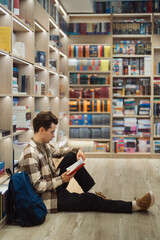 A young man sits on the floor in a well-lit, modern library, deeply engrossed in reading a book....