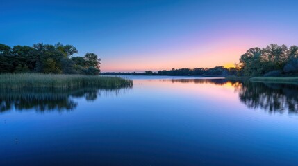 Fototapeta premium The image shows the tranquil waters of a lake reflecting the subtle colors of sunset with greenery framing the scene