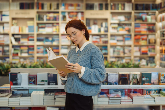A focused young woman in glasses and a blue sweater reads a book while standing in a vibrant, well-stocked bookstore. The shelves are filled with a variety of books.