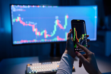 Oil prices moves up on mobile screen. Woman's hand holds a mobile phone. Monitor in the background.