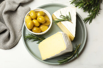 Plate with pieces of tasty camembert cheese, olives and rosemary on white table, top view