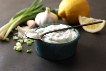 Delicious yogurt in bowl, green onion, garlic, lemon and spoon on black table, closeup