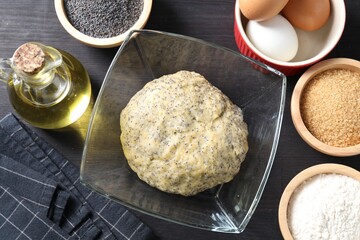 Raw dough with poppy seeds in bowl and ingredients on black wooden table, flat lay