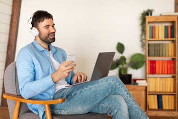 Smiling Man With Headphones Using Laptop and Holding Smartphone Indoors