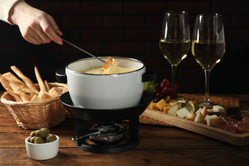 Woman dipping piece of bread into fondue pot with melted cheese at wooden table, closeup
