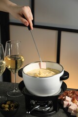 Woman dipping piece of ham into fondue pot with melted cheese at grey table with wine and snacks, closeup