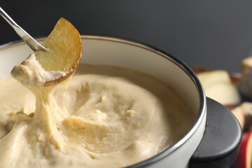 Dipping piece of potato into fondue pot with melted cheese on grey background, closeup