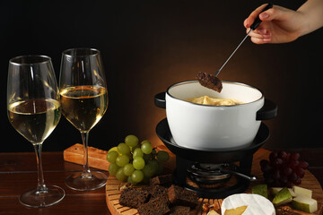Woman dipping piece of bread into fondue pot with melted cheese at wooden table with wine and snacks, closeup