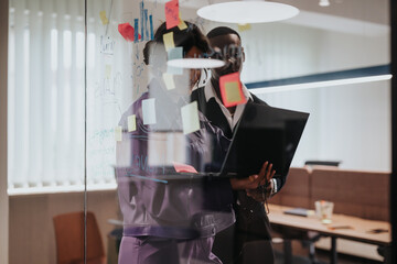 A focused African man and a Caucasian woman engage in a strategic planning session, using a laptop and sticky notes on a glass wall in a brightly lit office.