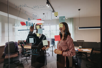 Energetic and creative brainstorming session of young, multiracial entrepreneurs using sticky notes and writing on glass walls in a modern office. - obrazy, fototapety, plakaty