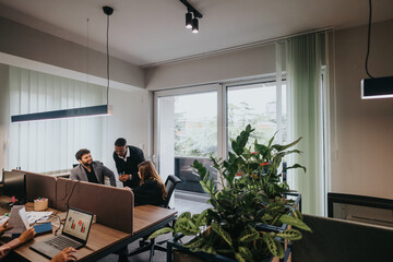 A group of diverse professionals collaborates around a workspace filled with natural light and plants in an indoor office environment.