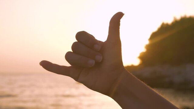 LENS FLARE, CLOSE UP, SILHOUETTE: Morning sunrise on beach and shaka hand gesture expressing good vibes and aloha spirit after fun surf session. Positivity and happiness on summer holidays at seaside.
