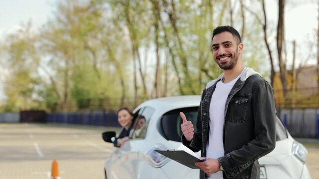 Arabic male driving instructor gives a thumbs-up sign as a gesture of passing a driver's test to a female student in a car behind him