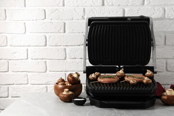 Electric grill with homemade sausages, rosemary and mushrooms on marble table. Space for text