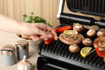 Woman cooking homemade sausages with mushrooms and bell pepper on electric grill at wooden table, closeup