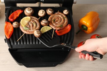Woman cooking homemade sausages with bell peppers and mushrooms on electric grill at wooden table, closeup
