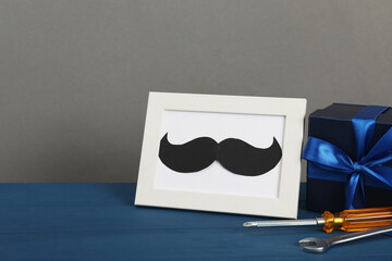Happy Father's Day. Gift box, tools and frame with mustache on blue wooden table, closeup. Space...