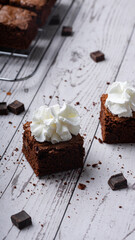 Chocolate chip brownies with whipped cream on a wooden background with crumbs and dark chocolate pieces around. Selective focus.