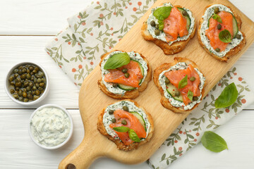 Tasty canapes with salmon, capers, cucumber and sauce on white wooden table, top view