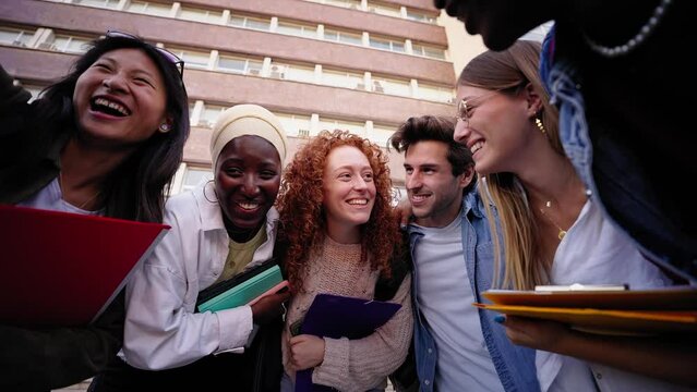 Low angle. Group of multiracial young cheerful students enjoying laughing outdoor. Diverse friends embracing with backpacks and workbooks gathered in circle having fun on a break of classes 