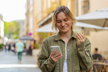 Young woman using credit bank card and smartphone while transferring money, purchases online shopping, order food delivery, booking hotel room. Happy girl walking on urban city sunny street outdoors.
