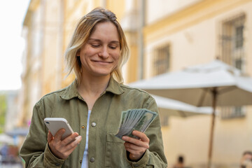 Happy young woman counting money dollar cash, using smartphone calculator app, satisfied of income for planned vacation gifts. Caucasian girl on urban city sunny street. Town lifestyles outdoors.