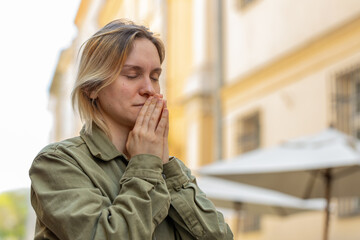 Portrait of young woman praying with closed eyes to God asking for blessing, help, forgiveness outdoors. Caucasian girl clasping hands wishing luck on sunny urban city street. Town religion lifestyles