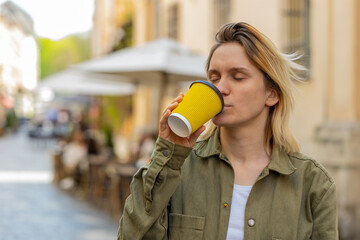 Happy Caucasian young woman enjoying morning coffee hot drink and smiling outdoors. Relaxing, taking a break. Girl walking through urban city center street, drinking coffee to go. Town lifestyles.