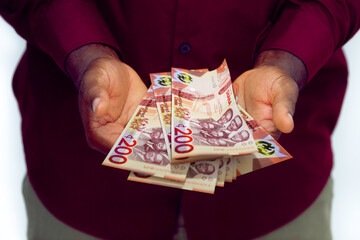 Black man in red long sleeve shirt holding Ghanaian cedi notes with opened palms

