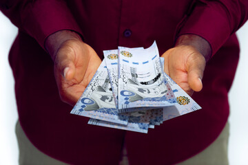Black man in red long sleeve shirt holding Saudi riyal notes with opened palms
